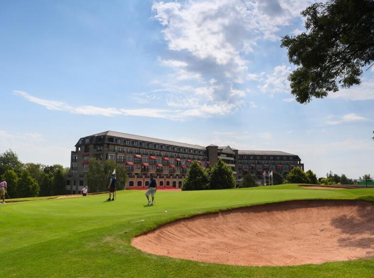 Golfers playing a round of golf by a bunker with a grand hotel in the foreground.
