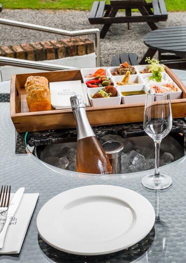 A selection of food in a wooden tray with a loaf of bread set on a table laid out with crockery and wine.