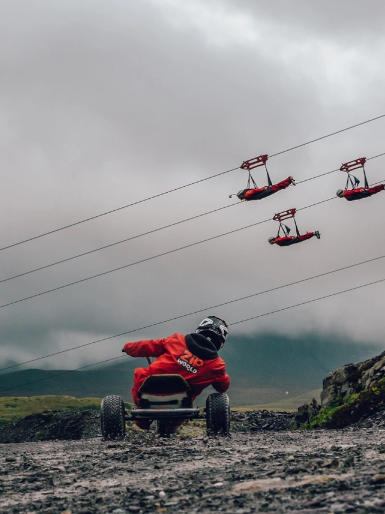 A go carter driving down a quarry with people passing by on zip lines.