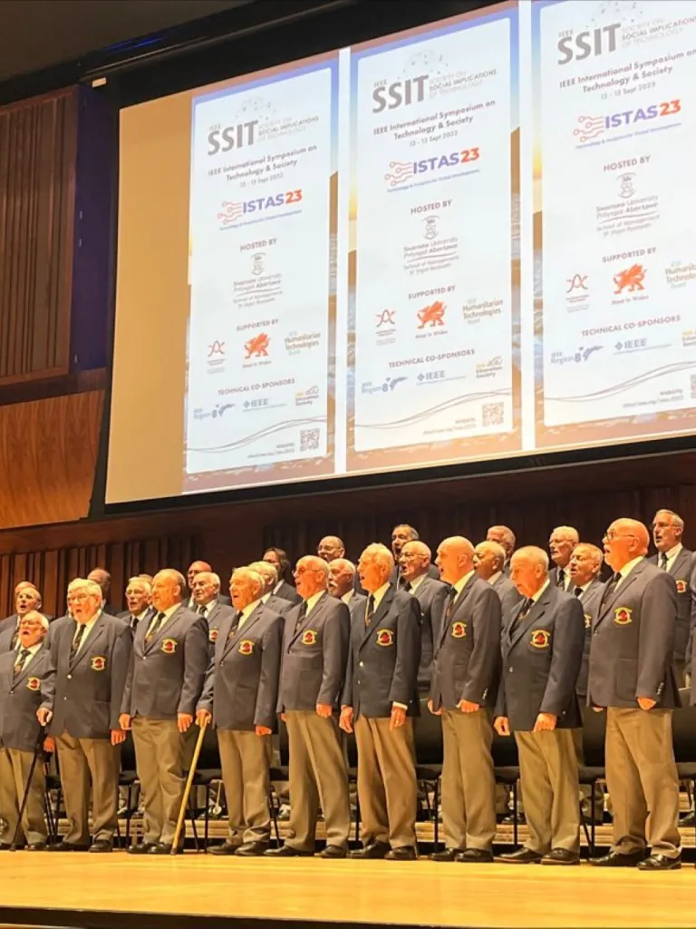 male voice choir on stage in suits with the conference banner behind them
