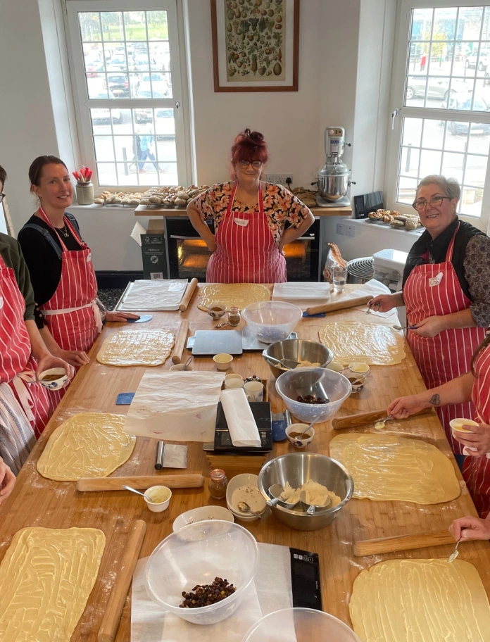 Abergavenny Baker Cookery School class smiling up at camera whilst wearing aprons and baking around a wooden table