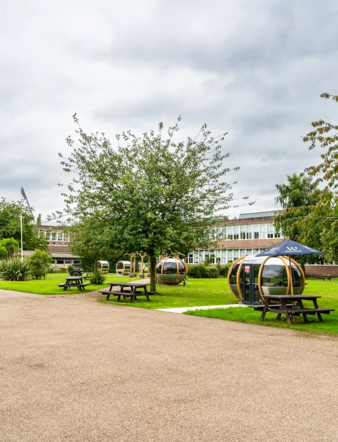 exterior shot of Wrexham University with green trees and gravel with seating and pods