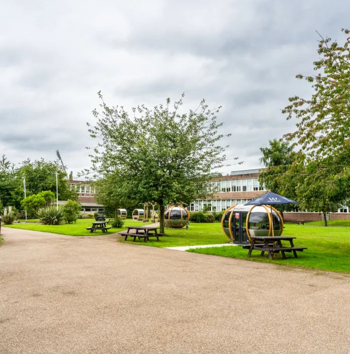 exterior shot of Wrexham University with green trees and gravel with seating and pods