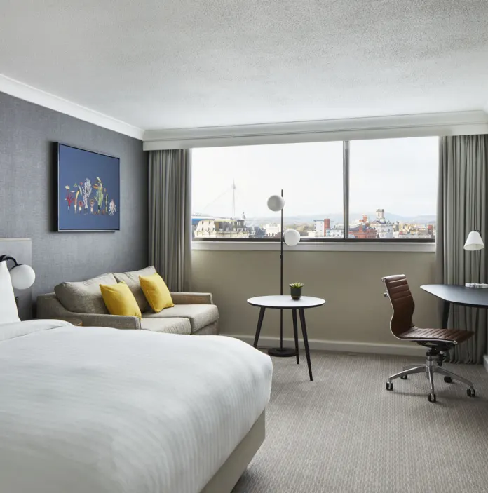 Bedroom featuring a plush bed, comfortable sofa with accent cushions, a workspace with city views through a large window, and contemporary décor creating a stylish, relaxing stay environment.