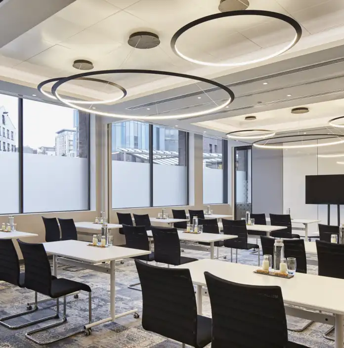Meeting room featuring modern tables and chairs, large windows with city views, and circular ceiling lights, set up for a professional corporate event or training session.
