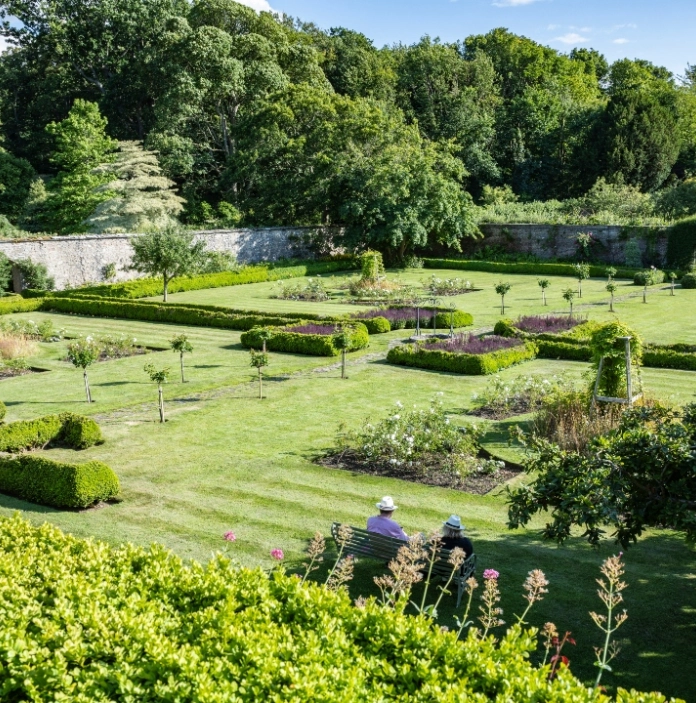 A formal garden with neatly trimmed hedges, flower beds, and two people sitting on a bench, surrounded by lush trees.