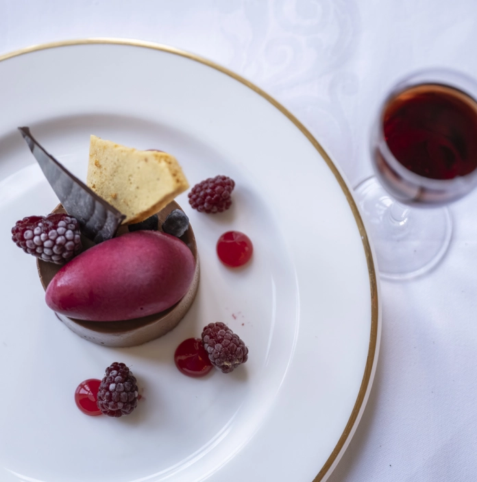 A plated chocolate dessert with berry sorbet, raspberries, and garnishes, served beside a glass of red dessert wine.