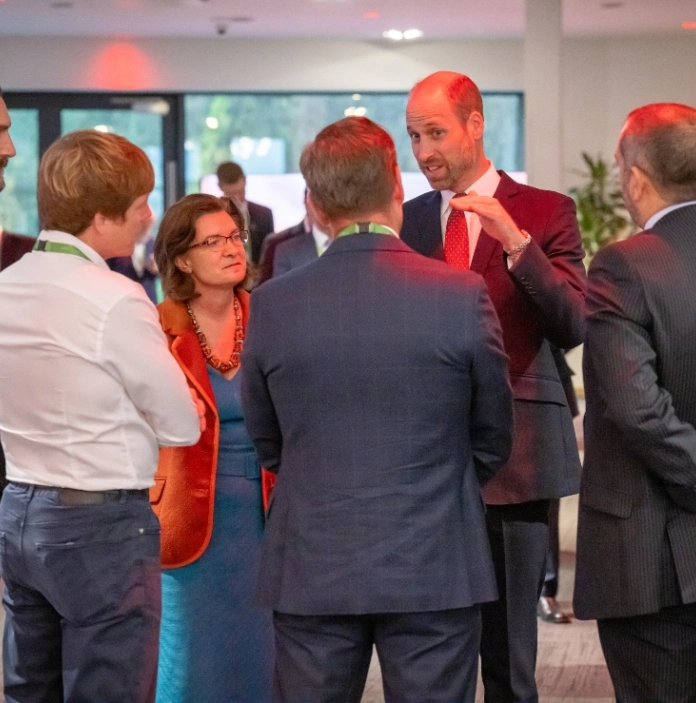  A group of senior attendees in formal attire stand in conversation at an indoor investment summit, with exhibition lighting and other delegates in the background.