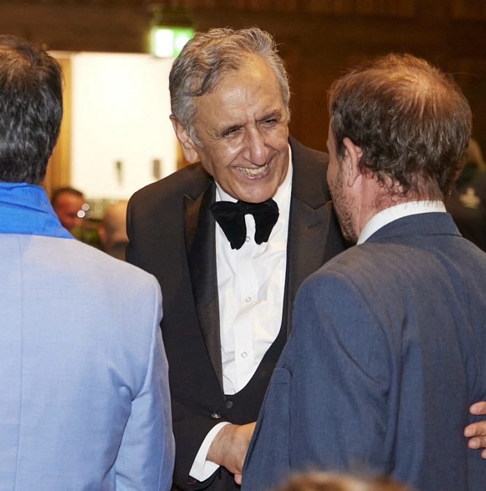 Three people in formal evening attire greet and shake hands at an indoor event.