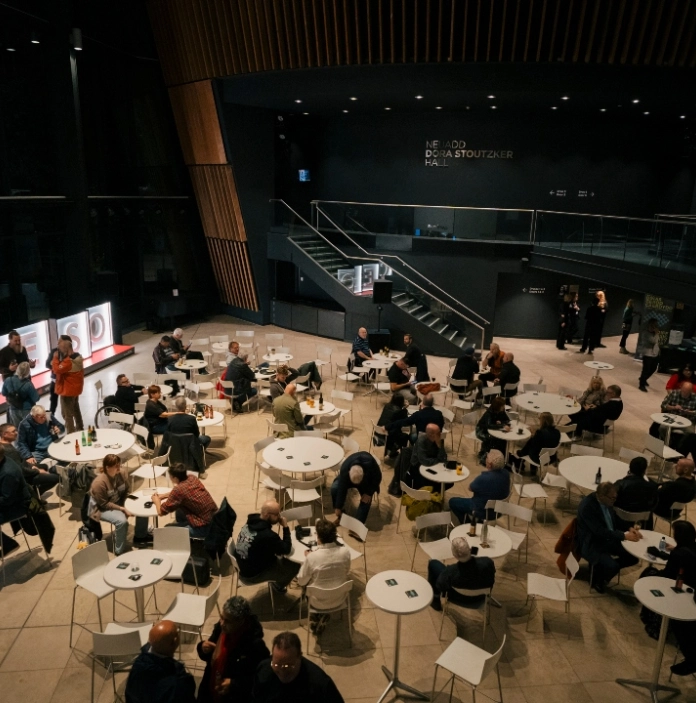 Crowd seated at café tables in a modern venue foyer with large “CROESO” sign and staircase