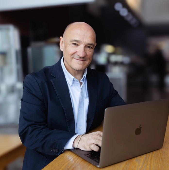 erson in a dark blazer sitting at a wooden table, using a laptop in a softly blurred café-style setting.