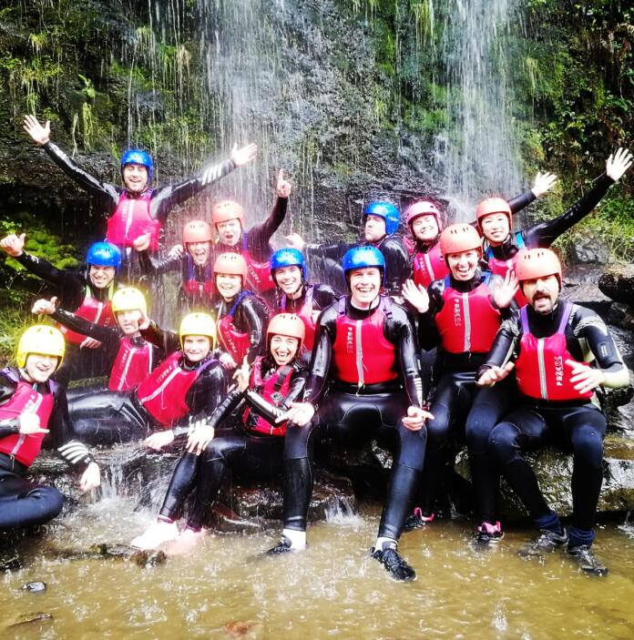 A group of people wearing wet suits, helmets and life jackets under a waterfall.