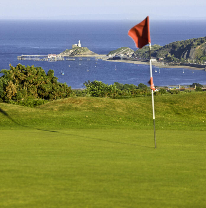 A flag in the hole on the green of a golf course with views of the bay beyond.