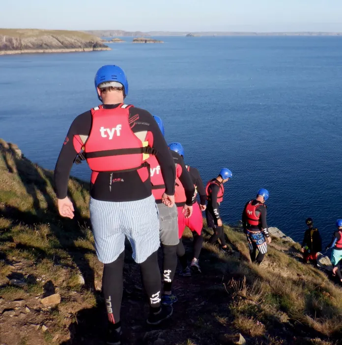 Coasteers wearing protective gear walking down a cliff toward the ocean.