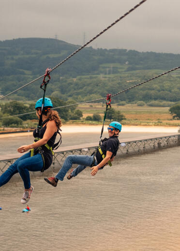 Two people zip wiring over a surfing lagoon with the countryside in the background.
