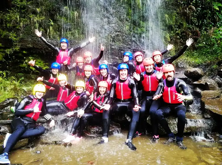 A group of people wearing wet suits, helmets and life jackets under a waterfall.