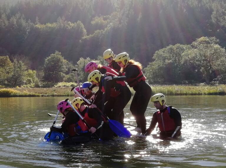 Group wearing safety gear climbing onto a blue raft in the water at a team building event.