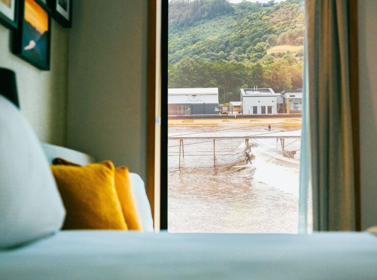 A bed with a view of surfers riding the waves outside the window.