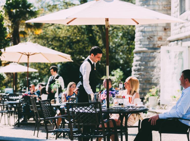 Waiters serving customers at outdoor tables covered with parasols in the sunshine.