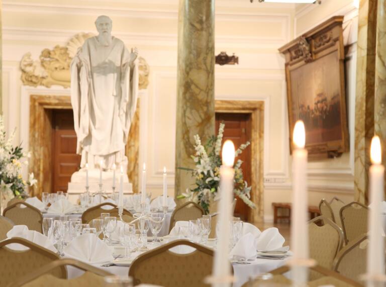 A table decorated with dinnerware and candles in a grand hall with pillars overlooked by a statue.