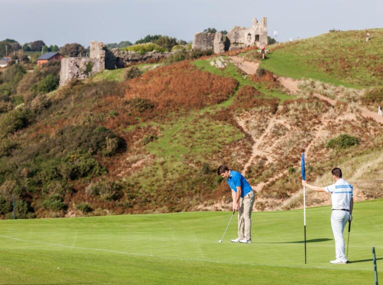 Two golfers on the green with the ruins of a castle behind on a gorse covered hill.