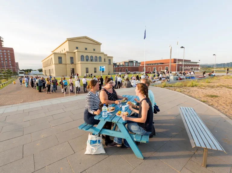 Delegates sitting on a picnic table enjoying a beach barbecue with the university in the background.