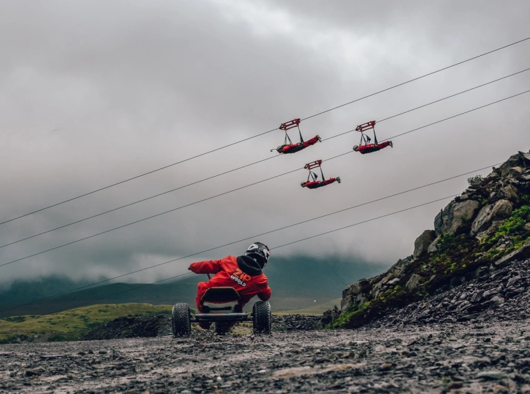 A go carter driving down a quarry with people passing by on zip lines.