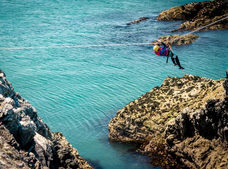 A person in safety gear traversing on a rope across the sea.