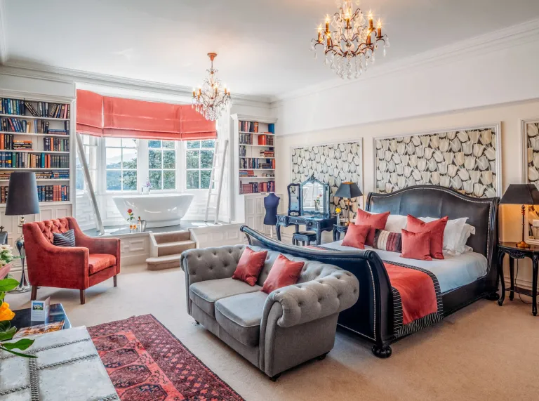orange and cream bedroom with large leather bed and orange pillows, cream carpet,grey settee and chandeliers.