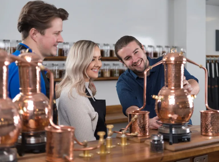 Woman with blonde hair and man with brown hair smiling , looking at copper gin distillery equipment and staff member presenting.