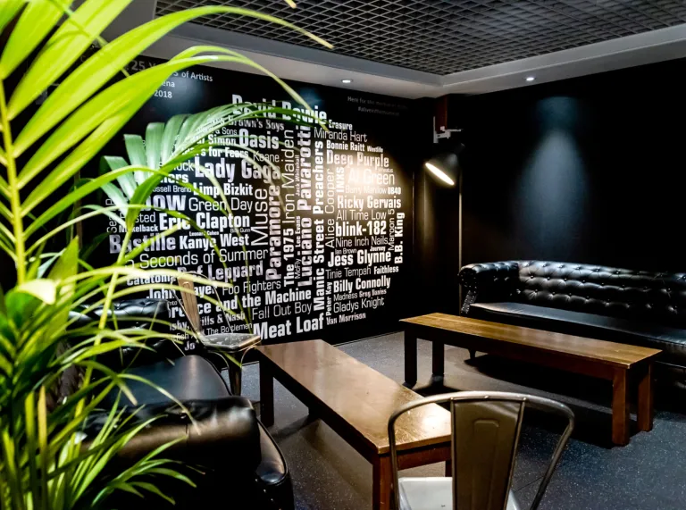 Interior room with black chesterfield settee, famous pop artist's names on walls, wooden benches and green leafy plant.