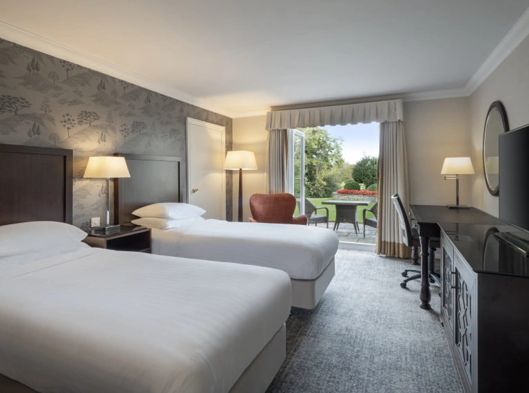 twin beds with white sheets and adesk and mirro with chair overlooking the patio doors onto the green grass and surrounding trees
