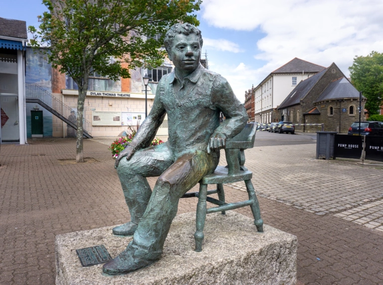 stone statue of Dylan Thomas sitting on stone with gardens behind