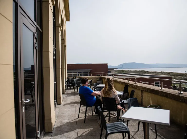Two people sitting on a balcony at the university looking out at the sea and beach