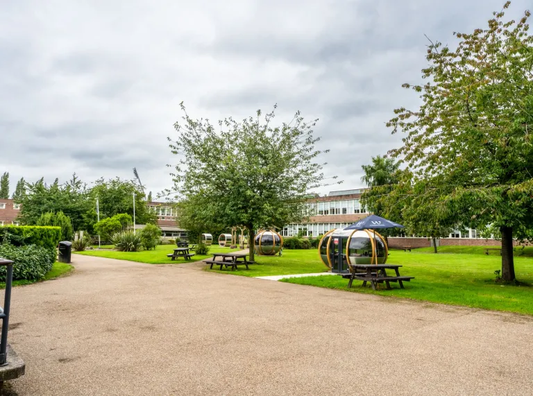 exterior shot of Wrexham University with green trees and gravel with seating and pods