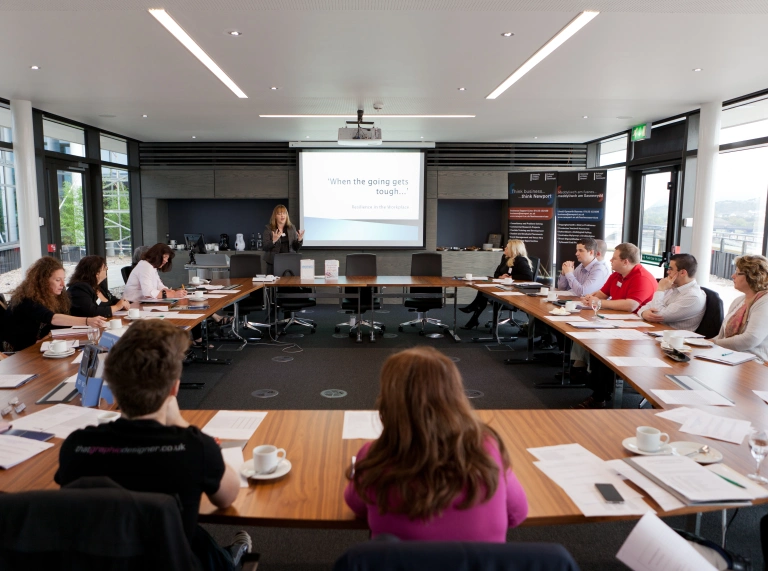 image of peopole sitting around a boardroom table listeningto someone talking and pointingat a whiteboard 