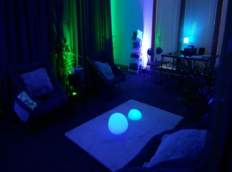 A dimly lit relaxation room with blue and green mood lighting, soft chairs, plants, and glowing orb lights on a rug