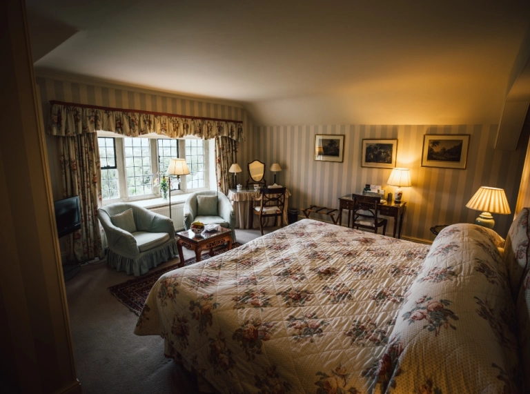 A warmly lit bedroom with a large floral bedspread, seating area by the window, and classic decor