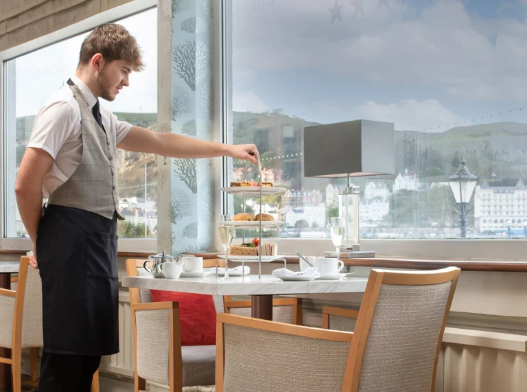 Staff member serving afternoon tea beside a window with a seaside town view
