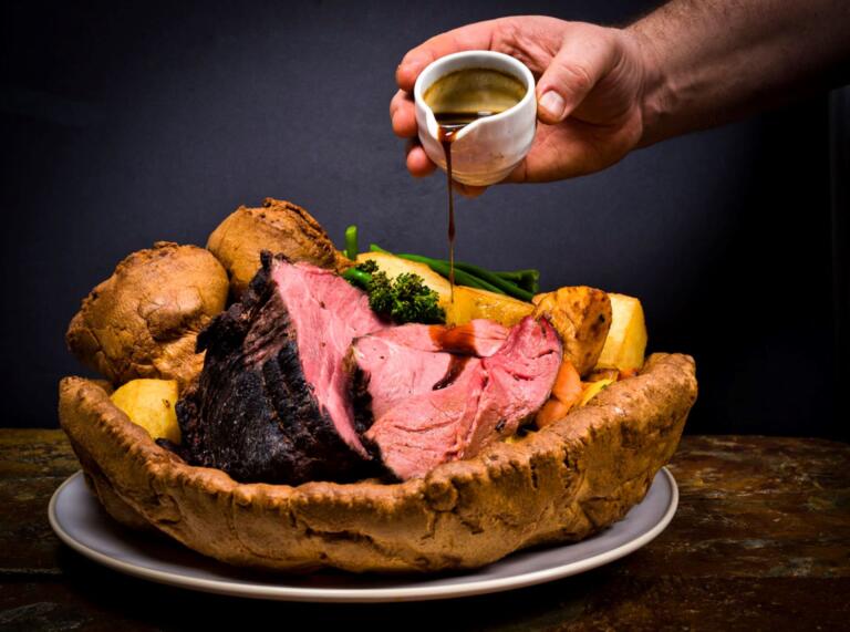 A jug of gravy being poured over meat, potatoes and vegetables in a yorkshire pudding.