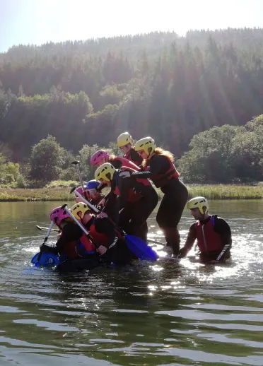 Group wearing safety gear climbing onto a blue raft in the water at a team building event.