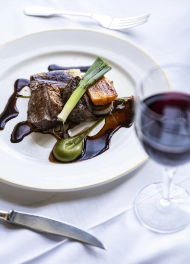 A plated fine-dining dish with braised meat, vegetables, and sauces, served alongside a glass of red wine.