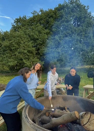 Group gathered around a large outdoor fire pit roasting marshmallows on sticks, with wooden benches and lush green trees in the background under a clear sky