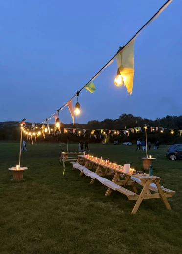 Outdoor evening setup at Garth Glamping with long wooden picnic tables arranged under string lights and bunting, surrounded by open grassy fields and distant trees under a dusky blue sky