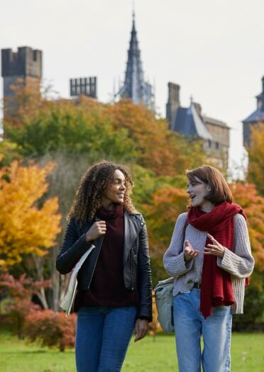 Two ladies walking through a park with golden leaves on the trees and a castle in the background.