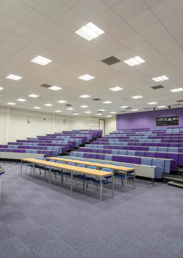 A lecture theatre showing the rising rows of purple furnished chairs and carpet.