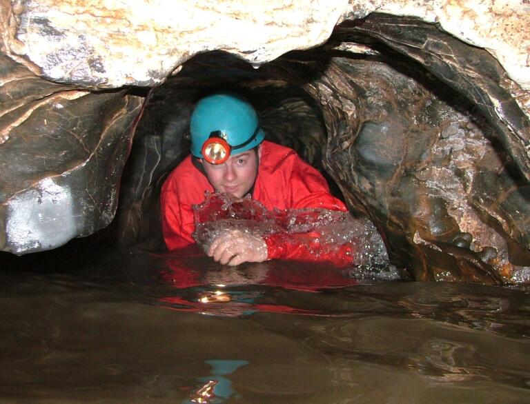 A man wearing a safety hat with a lamp coming out of a cave filled with water.