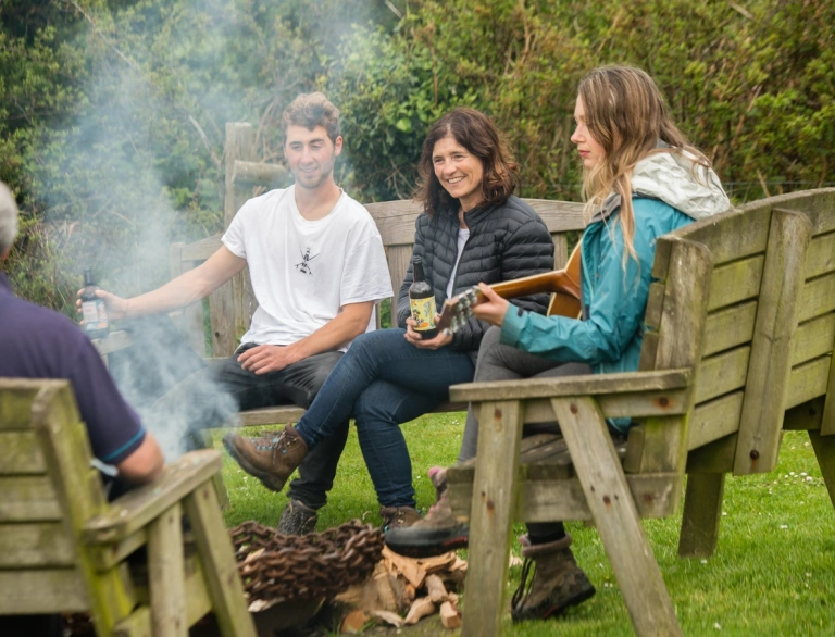 People sitting on benches around a campfire, with a guitar and drinks.