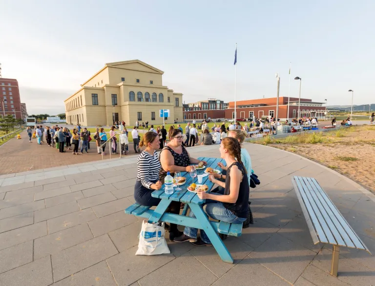 Delegates sitting on a picnic table enjoying a beach barbecue with the university in the background.