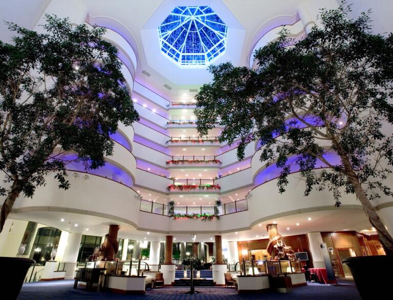 A large atrium in a luxury hotel with symmetrical trees and a glass dome roof. 